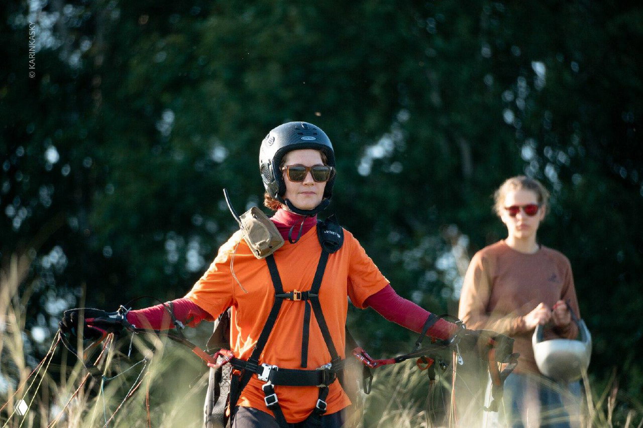 Пилот в шлеме и страховочной системе идёт по высокой траве с парапланом, подготовка к старту на фоне деревьев и помощника.