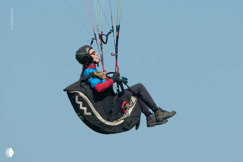 Пилот в подвесной системе парит в небе, виден профиль крыла параплана на фоне чистого голубого неба.