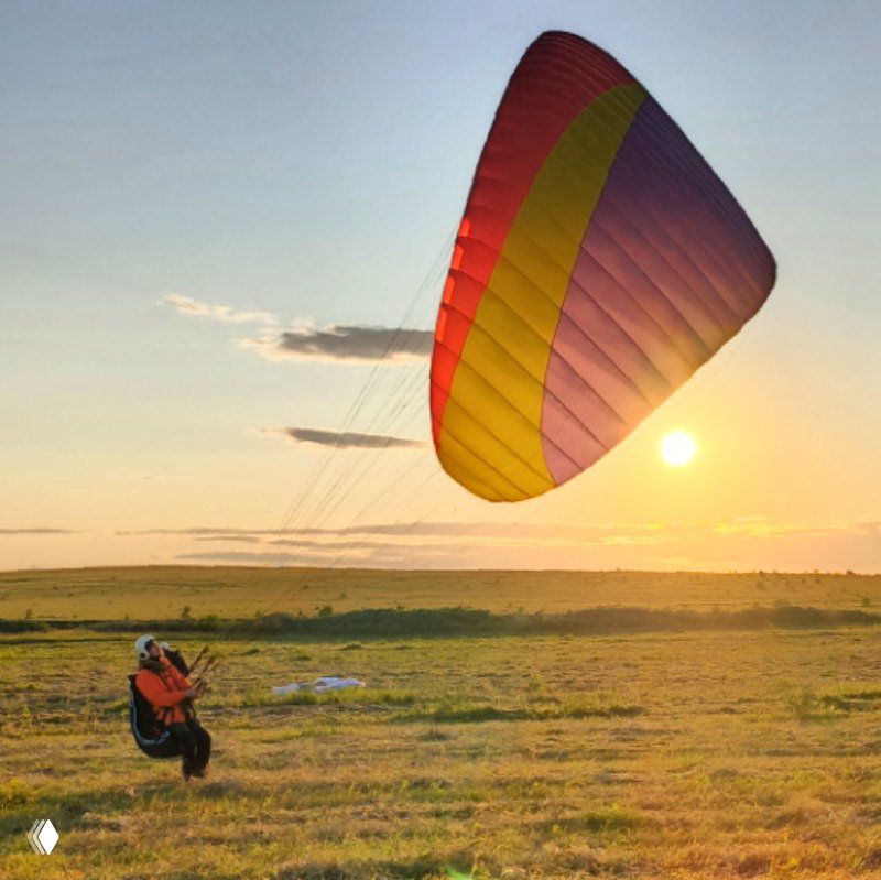 Купол параплана на закате: пилот готовится к взлёту на фоне тёплого солнечного света и широкого поля перед заходом солнца.