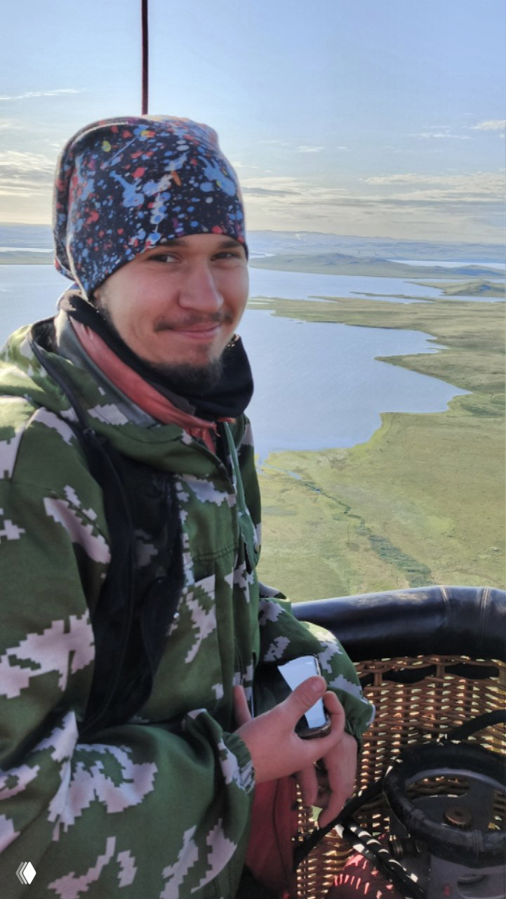 Пилот в корзине воздушного шара над Хакасией: улыбка в кадре, под ним видны озёра и равнины в мягком вечернем освещении.