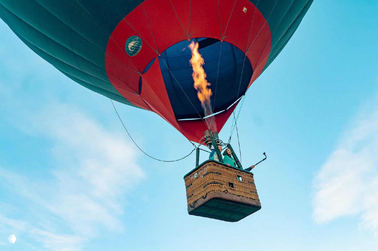Купол яркого аэростата и пламя горелки в момент подъёма: корзина с людьми под чистым голубым небом, вид снизу.