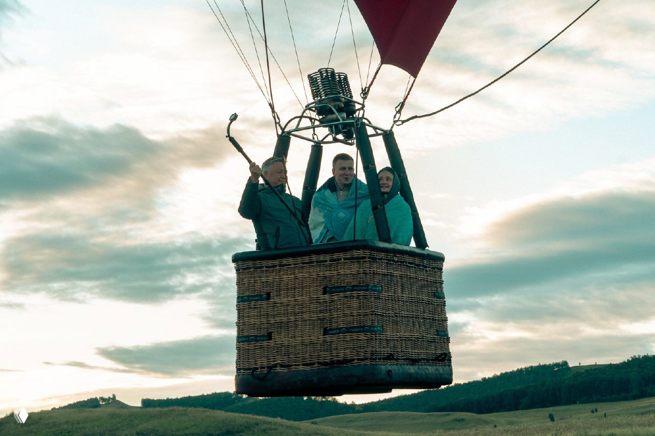 Крупный план плетёной корзины воздушного шара с тремя пассажирами и оборудованием горелки на фоне облачного вечера и зелёного луга.