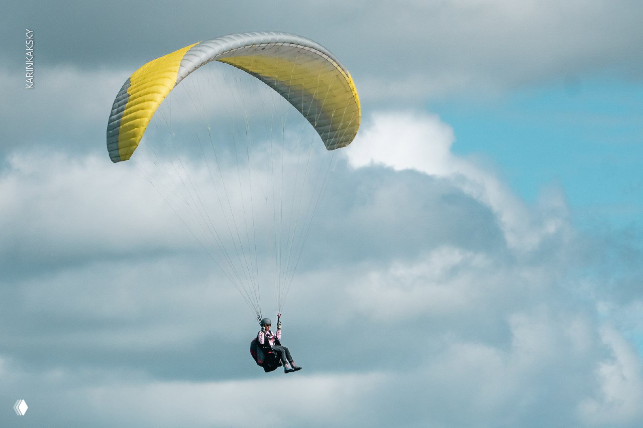 Пилот на жёлтом параплане в высоком ракурсе на фоне объёмных облаков — кадр показывает плавность полёта и положение пилота.