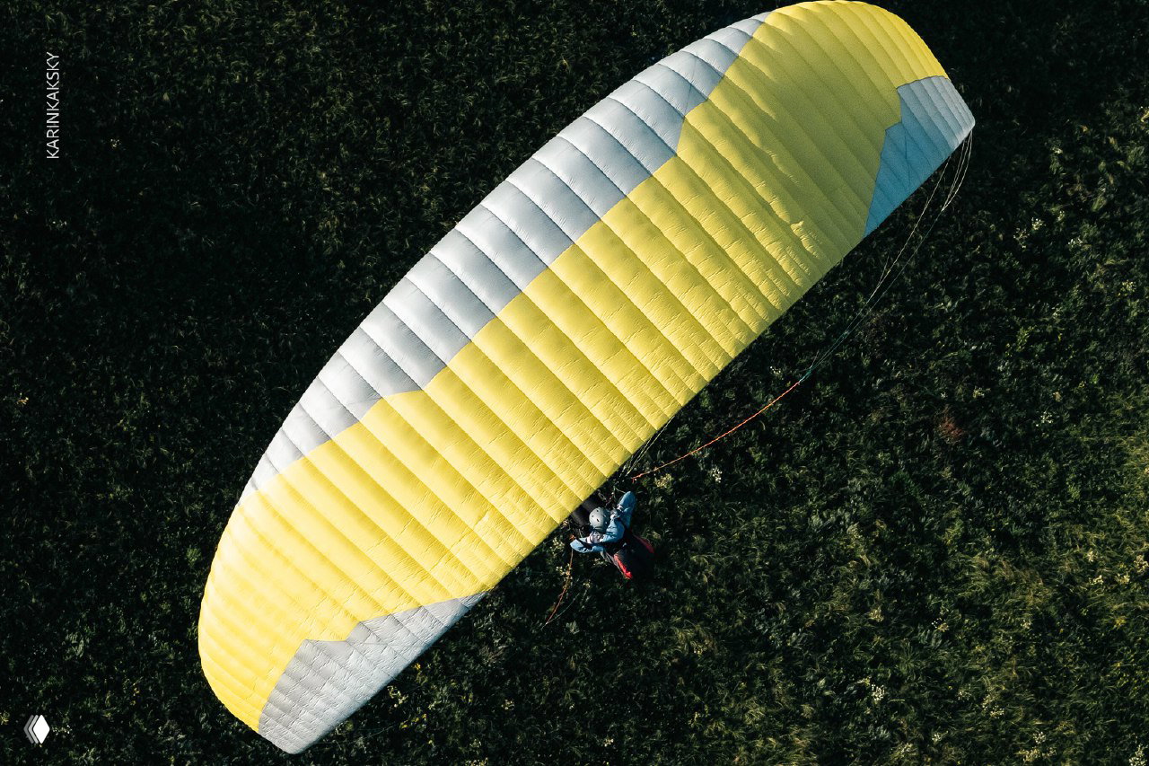 Крупный вид крыла параплана жёлто‑серой расцветки сверху над зелёной травой, видна текстура ткани и распределение ячеек крыла.