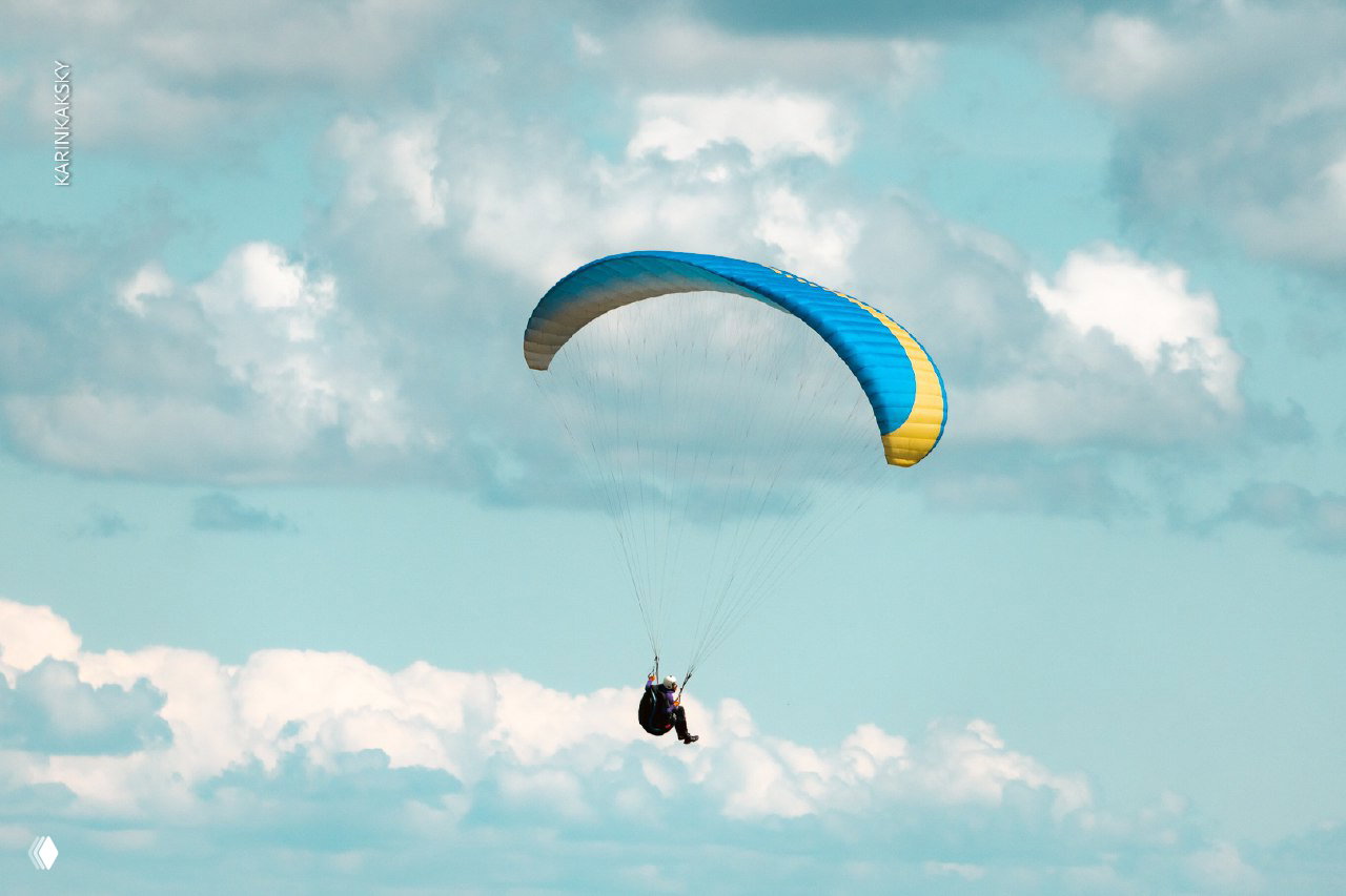 Одинокий пилот на параплане на фоне голубого неба и кучевых облаков, вид снизу и акцент на крыле синего и жёлтого цветов на фоне неба.
