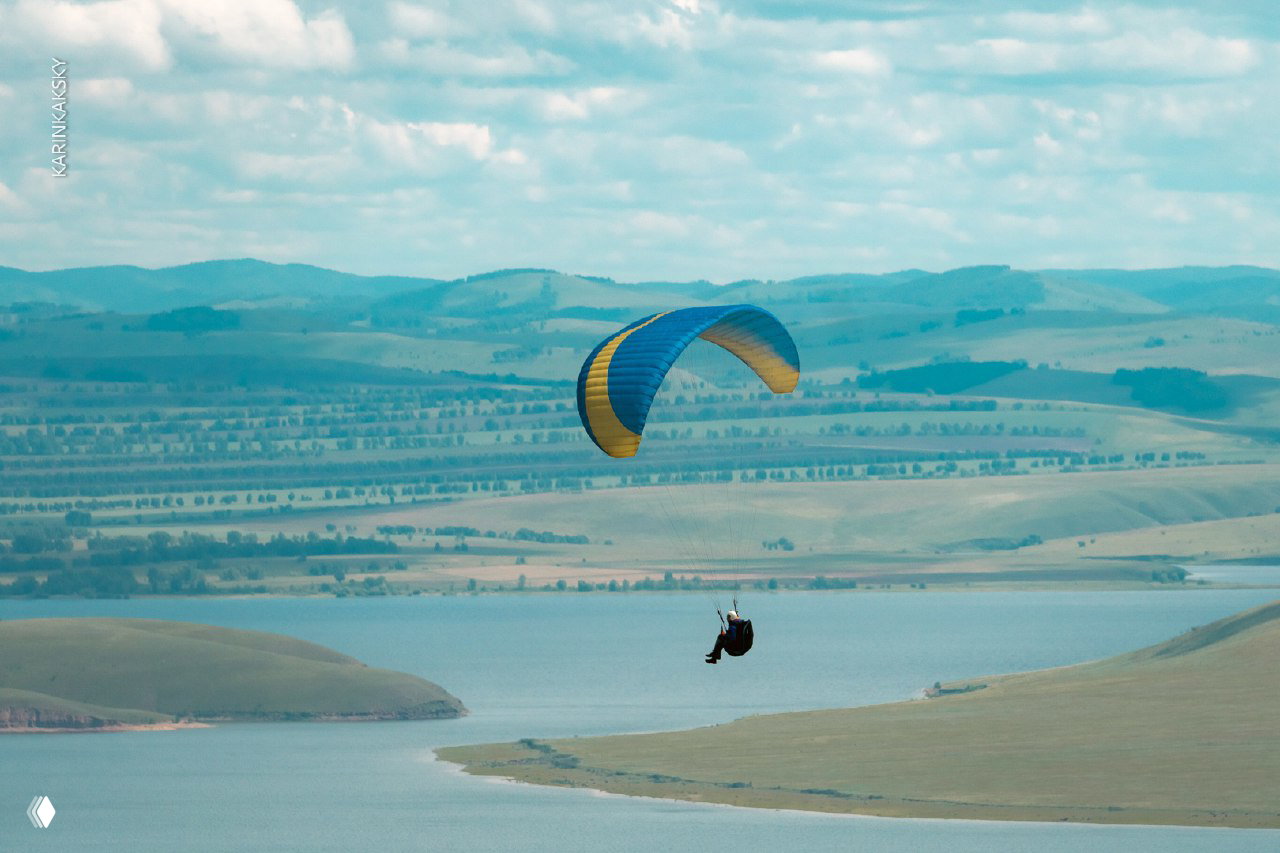 Парапланист над береговой линией и водной гладью, в кадре просматриваются поля и линии холмов, спокойная летняя сцена в воздухе.