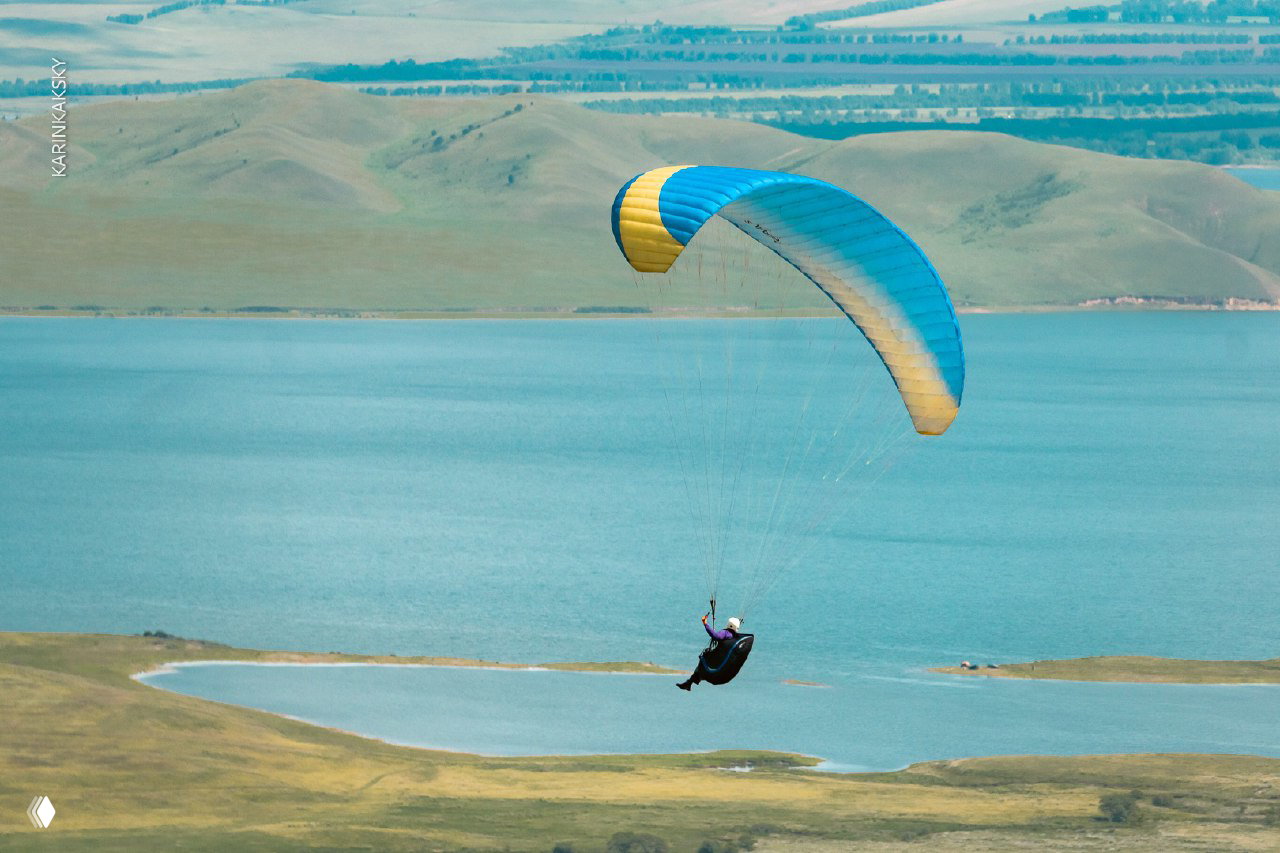 Крупный план параплана с сине‑жёлтым куполом над голубым водоёмом, пилот в подвесной системе управляет крылом, широкая природная панорама.