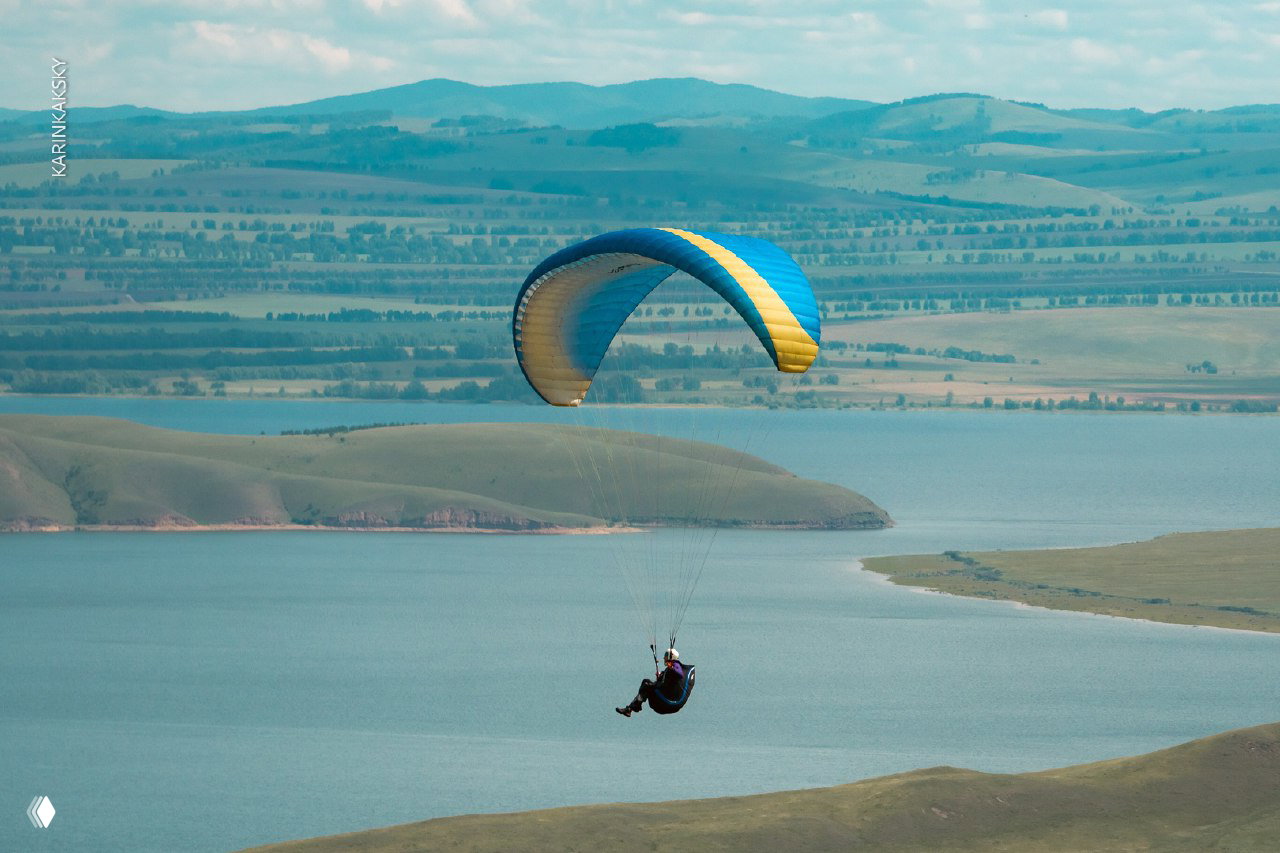 Парапланист летит низко над водной поверхностью между пологими холмами, спокойный летний пейзаж и широкий вид вокруг маршрута полёта.