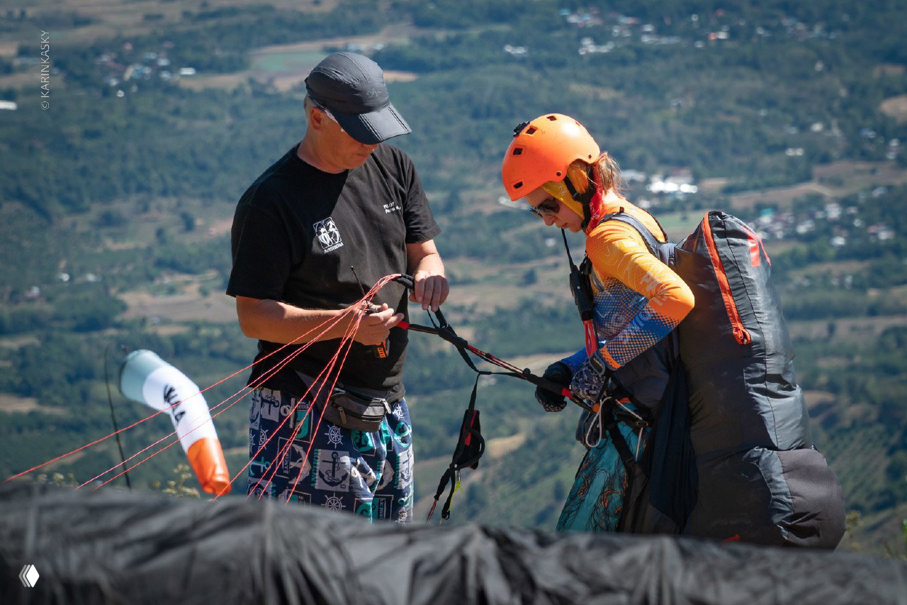 Инструктор проверяет снаряжение и стропы у пилота перед стартом на горном старте с видом на долину и деревни внизу.