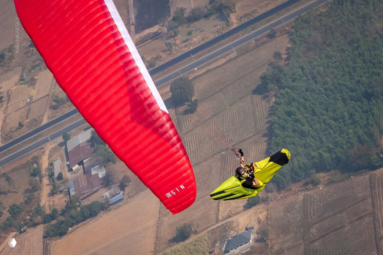 Пилот в желто-зелёном обтекателе и красное крыло параплана над сельской местностью, съемка сверху во время полёта.
