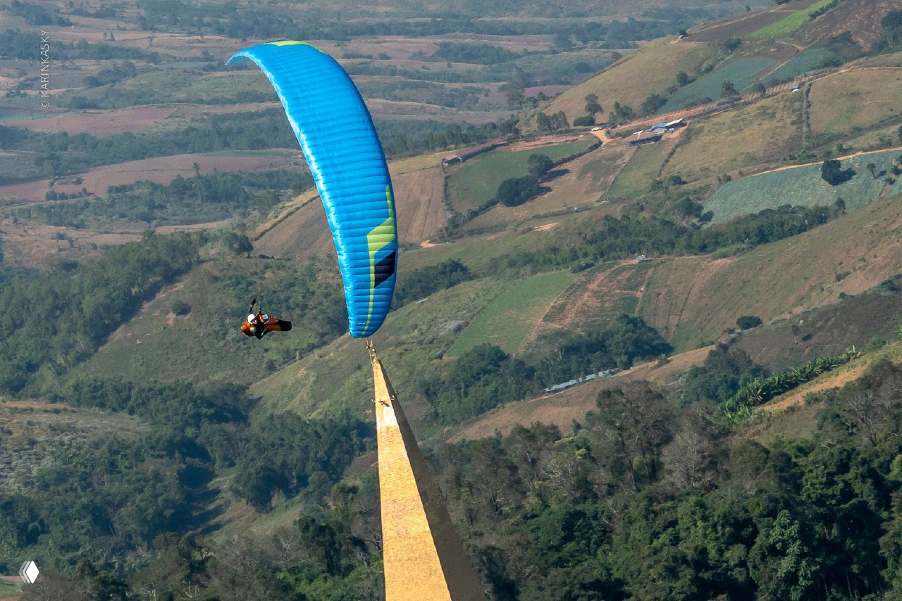 Синий параплан пролетает над острой скальной вершиной и зелёными холмами, демонстрируя типичные термические условия Тайланда зимой.