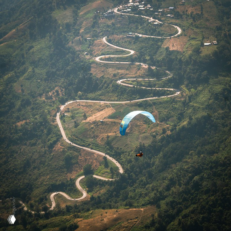 Извилистая горная дорога и одинокий парапланист над зелёными склонами, показывающие маршруты и ландшафтные особенности полётов.