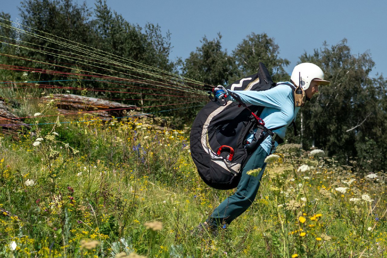 Динамический кадр: пилот делает беговой толчок по травяному склону, стропы натянуты, пейзаж летний и цветущий на переднем плане.