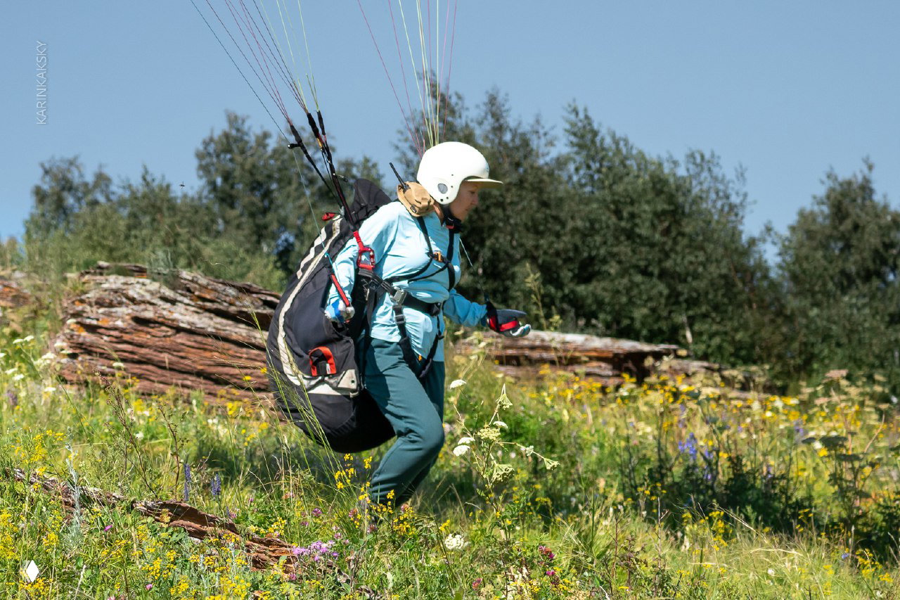 Пилот в гамаке уже частично оторван от земли, видна подушка подвески и береговая растительность; спокойная летняя погода и ясное небо.