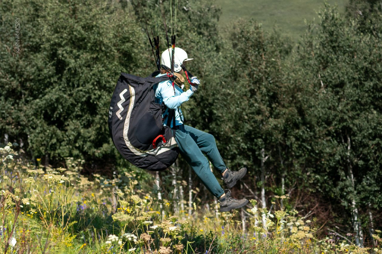 Пилот в полёте на фоне лесистого склона, вид на подвесную систему и стропы, ноги свободно виснут над лугом с цветами и зеленью.