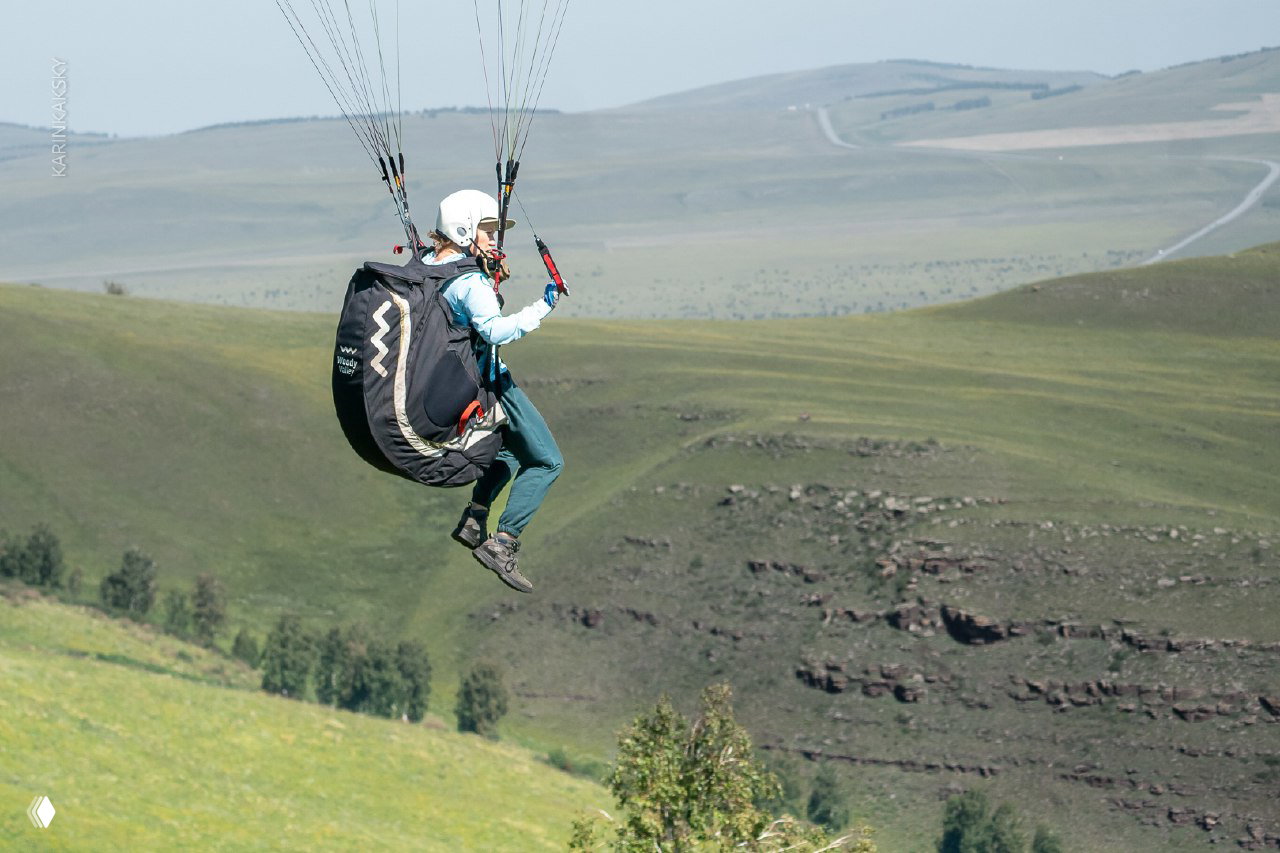 Небольшая высота: пилот висит в подвеске над цветущим склоном, вдалеке виден рельеф и деревья, ощущение тихого парения.