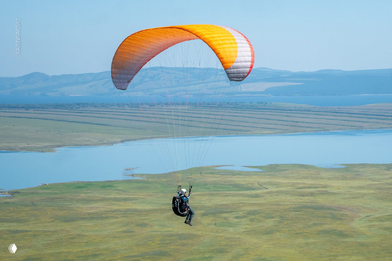Пилот на высоте, над широким холмистым ландшафтом Хакасии, видна подвеска и купол параплана, простор и озёрный фон на горизонте.