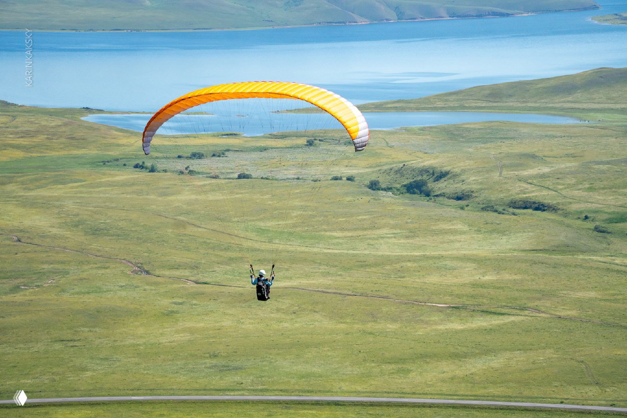 Широкий план: пилот в подвеске летит над зелёной равниной Хакасии, видно изгибы дорог и просторное небо — кадр подчёркивает свободу полёта.