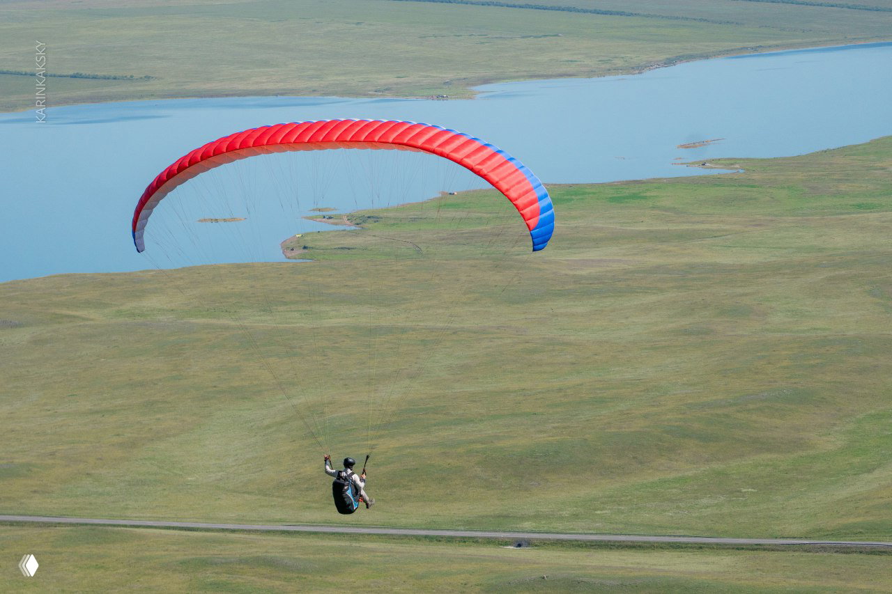 Парапланерист на красно-синем крыле парит над травяной степью у озера — вид сверху с майского или июльского паракэмпа в Хакасии.