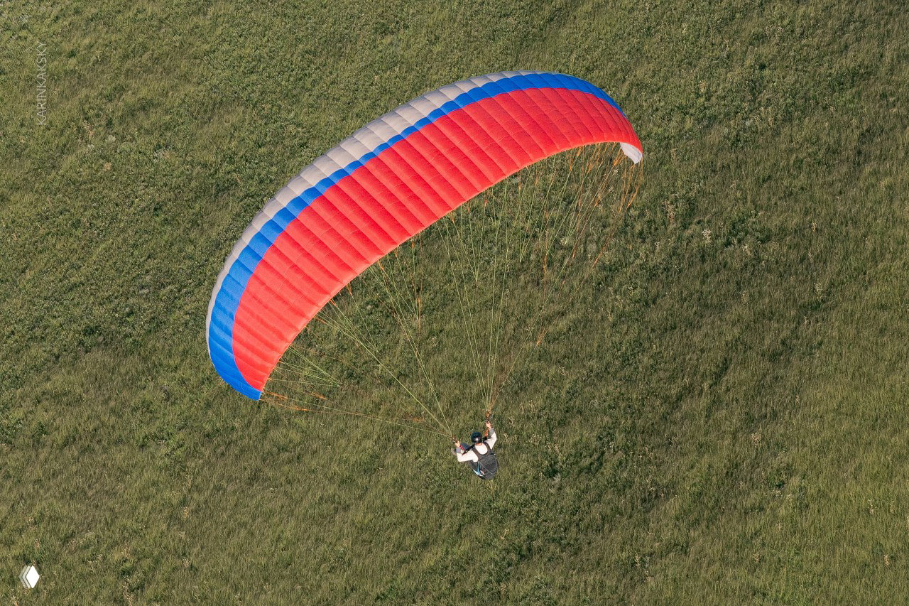 Вид сверху на крупное красно-синее параплан‑крыло и пилота над травой — детали строп и крыла, съёмка с паракэмпа в Хакасии.