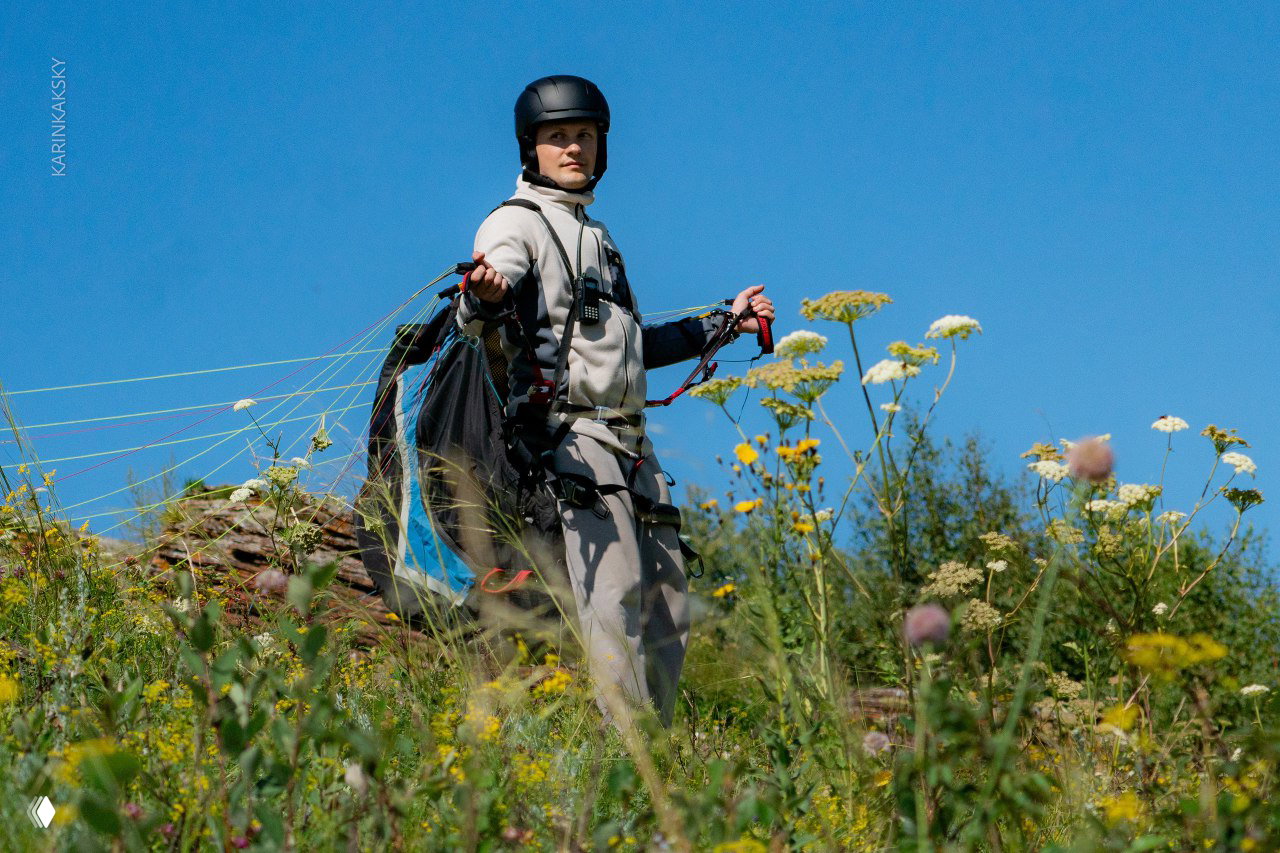 Пилот в шлеме и подвесной системе стоит на вершине среди летних цветов и трав, готовясь к взлёту на параплане — сцена с паракэмпа в Хакасии.