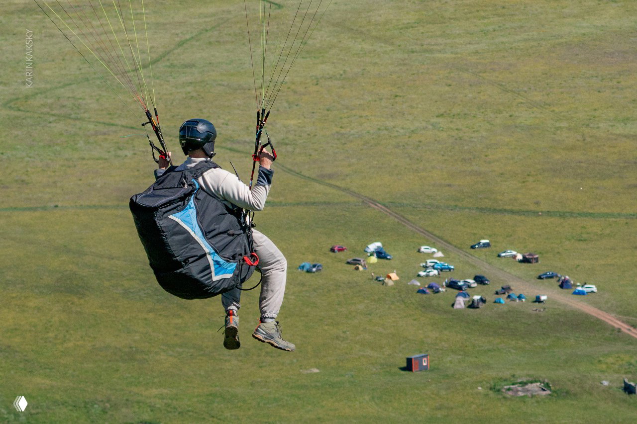 Парапланерист над палаточным лагерем и машинами на лугу — общий план с маршрута паракэмпа в Хакасии, вид с воздуха на базу выезда.