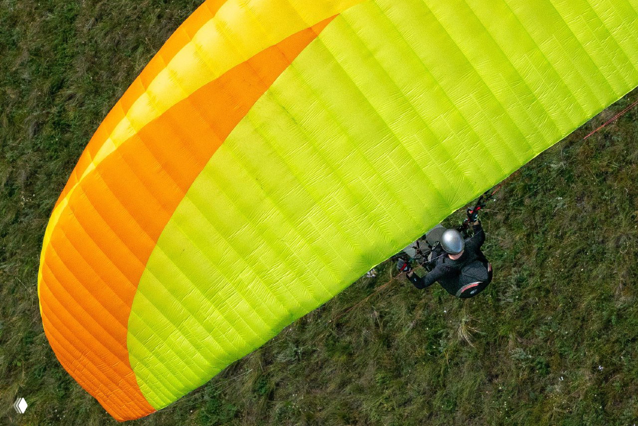 Крупный план ярко‑жёлтого и оранжевого купола параплана и пилота над травянистым склоном; видны стропы и элементы подвесной системы.