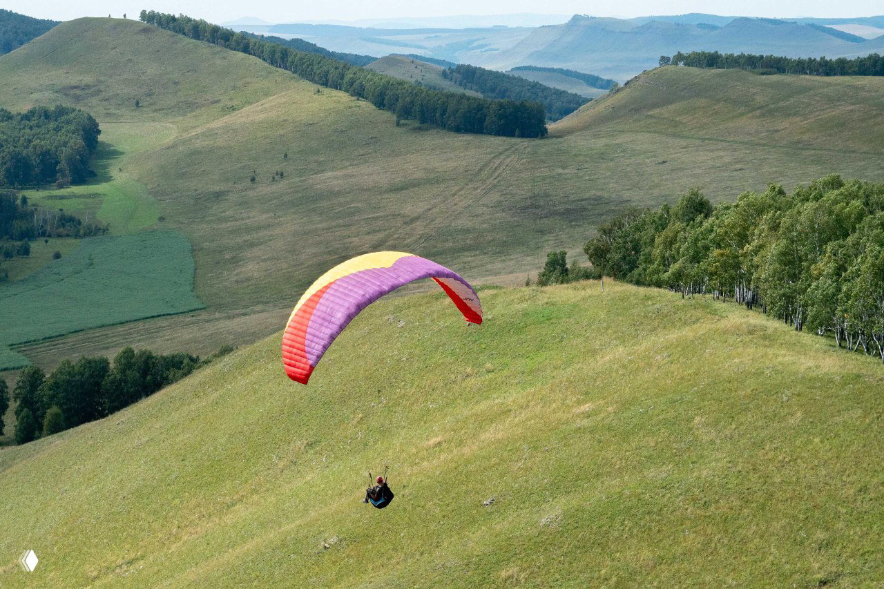 Пилот на параплане над зелёными холмами и лесной полосой в Аешке; видна широкая долина и учебные участки маршрута.