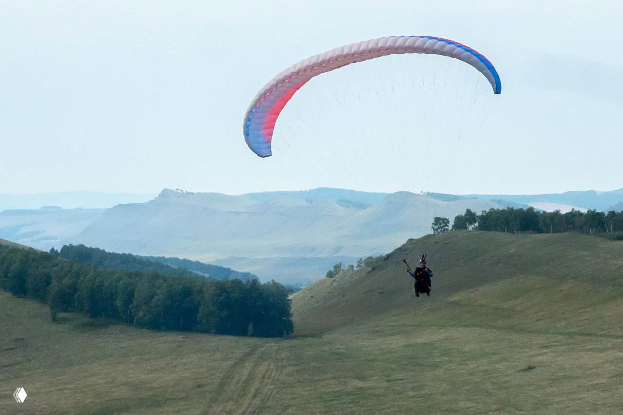 Парапланист в полёте над дальними хребтами и пастбищными склонами; купол пастельных тонов и спокойный пейзаж учебной зоны.