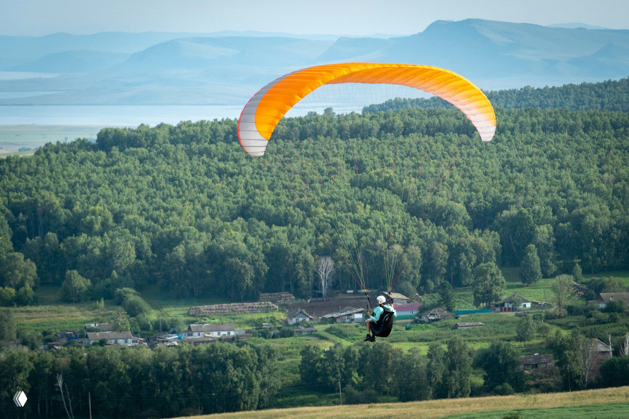 Параплан над лесистым склоном и небольшой деревней в Хакасии: оранжевое крыло на фоне леса, полей и водной глади.