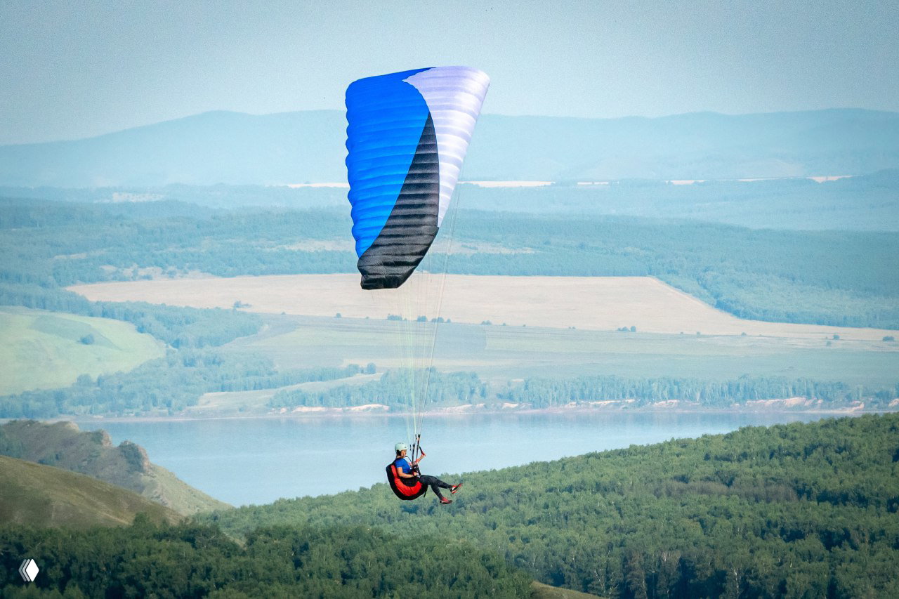 Парапланерист под сине‑белым крылом на фоне дальних холмов и реки — просторный летный пейзаж Хакасии с горизонтом.