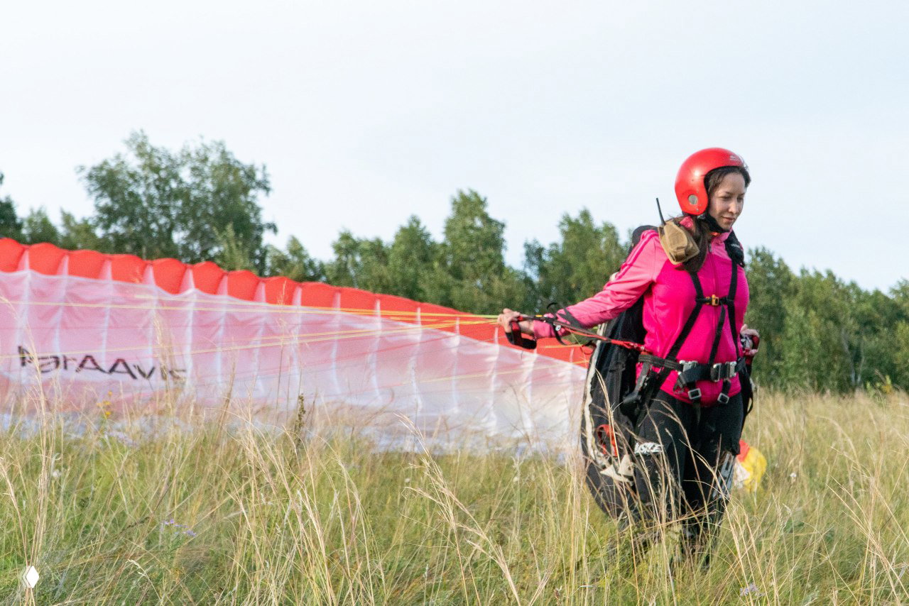 Подготовка к полёту: девушка в красном шлеме и страховке идёт по траве рядом с растянутым парапланом — момент из наземной съёмки.