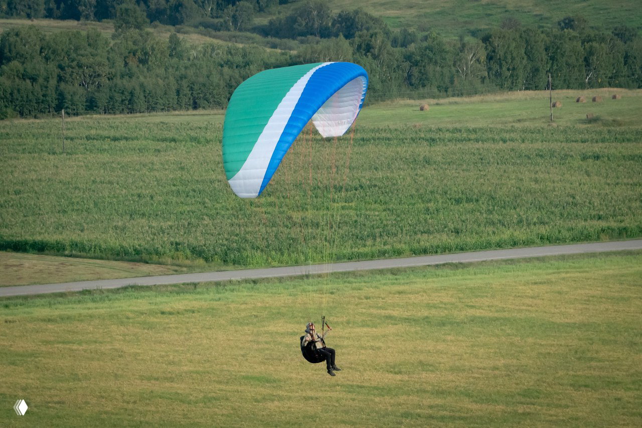 Парапланерист на зелёном крыле летит низко над сельским полем и дорогой в Хакасии — вид приближенного приземления и ландшафта.