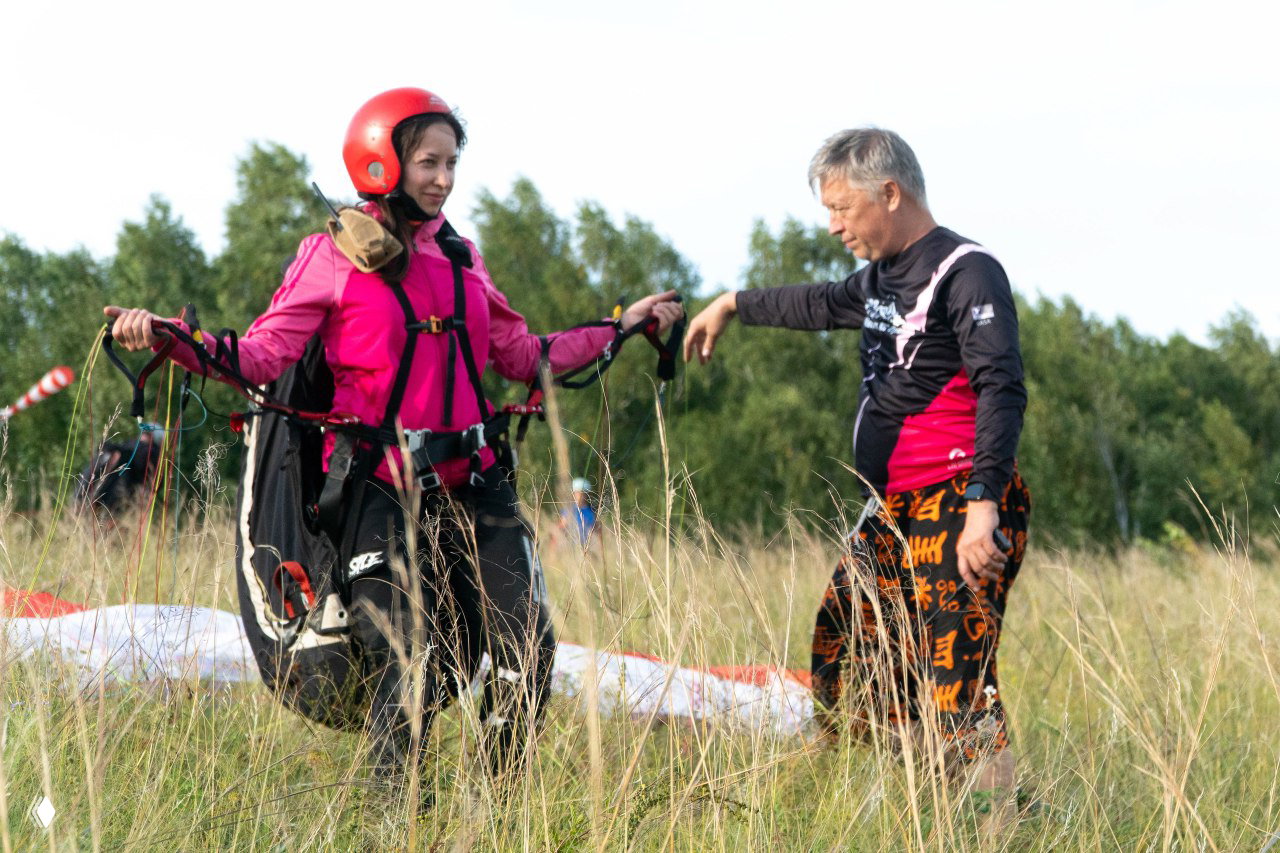 Ученица в розовом шлеме и инструктор обсуждают положение: жилет, обвязка и управление стропами перед взлётом на склоне.