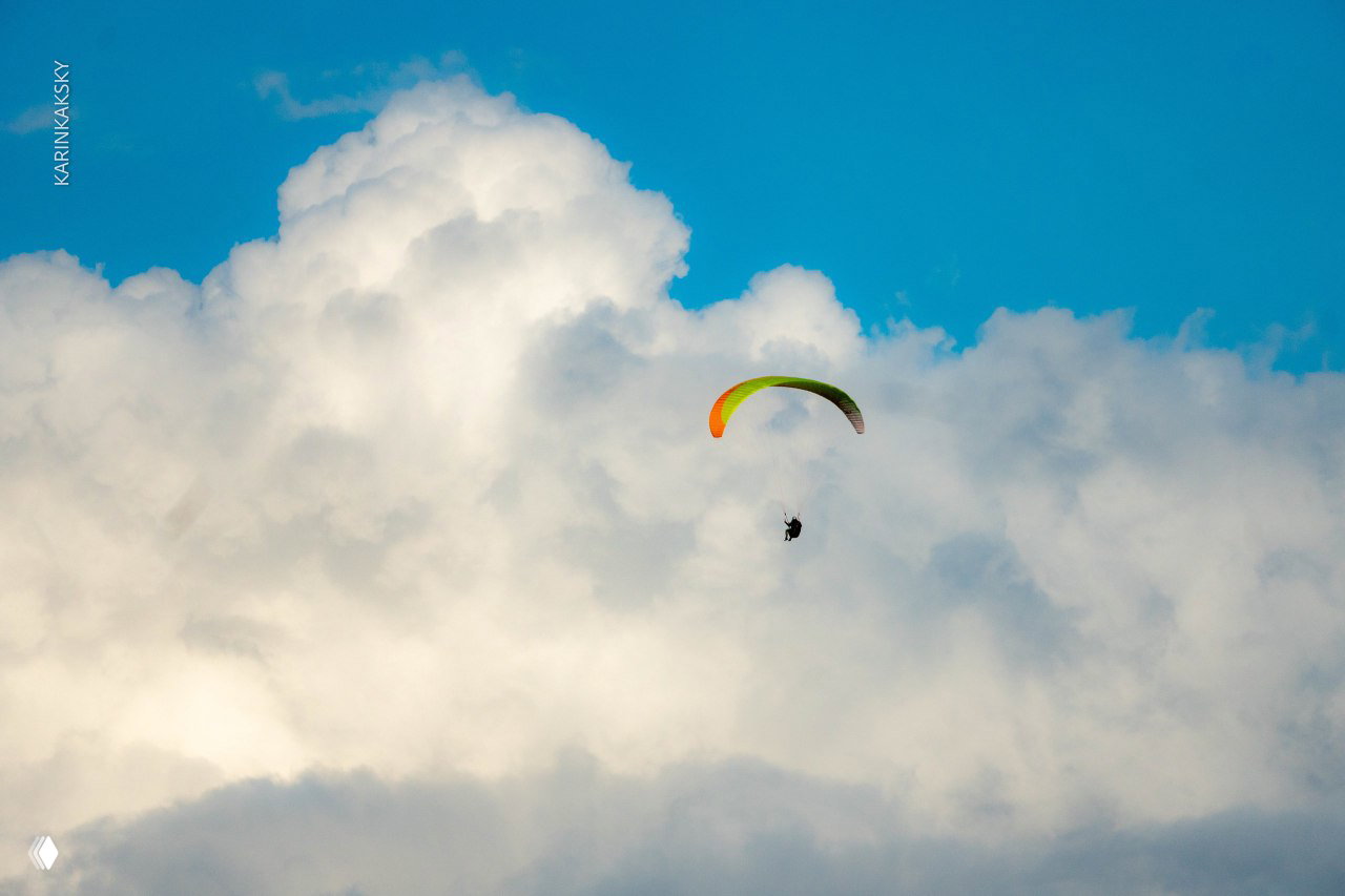 Параплан на фоне крупного кучевого облака и голубого неба: пилот в подвеске летит у облачного фронта, эффект масштаба.