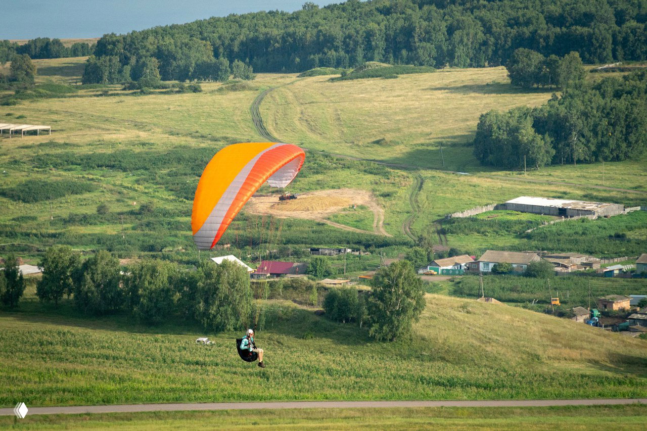 Параплан с оранжевым крылом над полями и деревнями: посадка и панорама холмов, видна сельская местность под крылом.