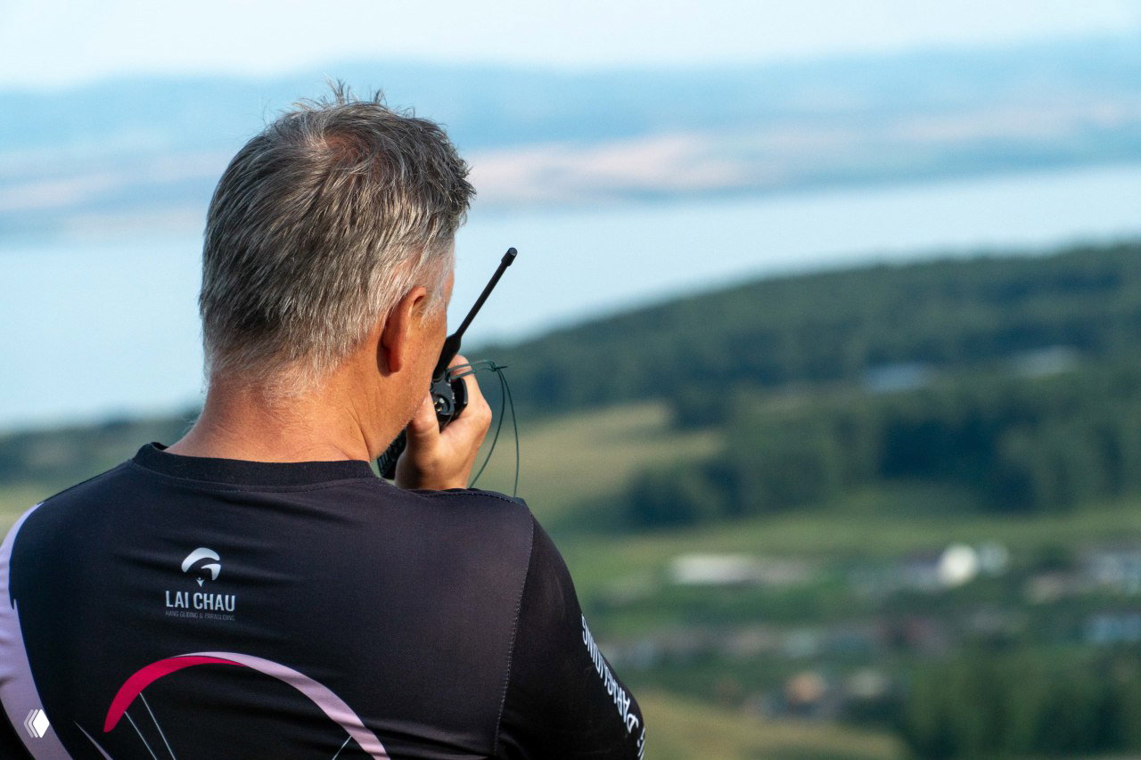Инструктор с рацией наблюдает за полётами с вершины склона: обратный вид на ландшафт, озеро и удалённые поля за ним.