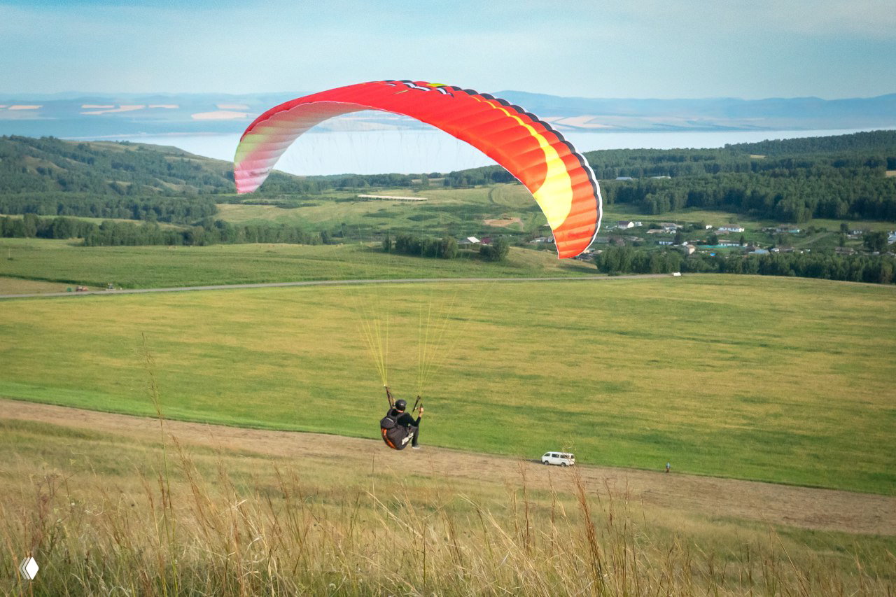 Пилот на параплане с ярко-красным крылом летит над холмом; широкая зелёная долина, дороги и сельские постройки видны внизу.