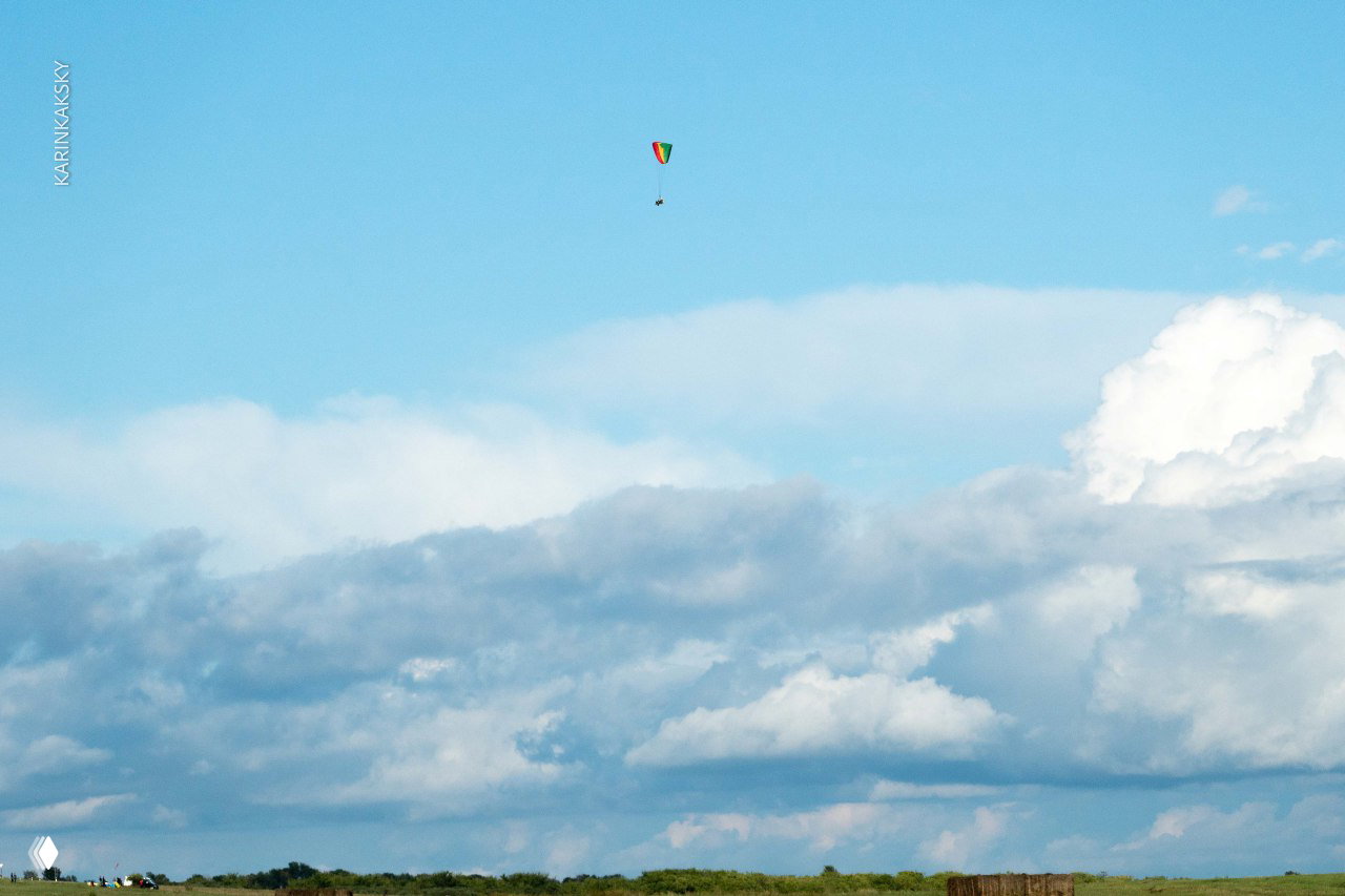 Маленький разноцветный параплан высоко в ясном небе над плотным слоем облаков; ощущение высоты и свободы, удалённый масштаб.