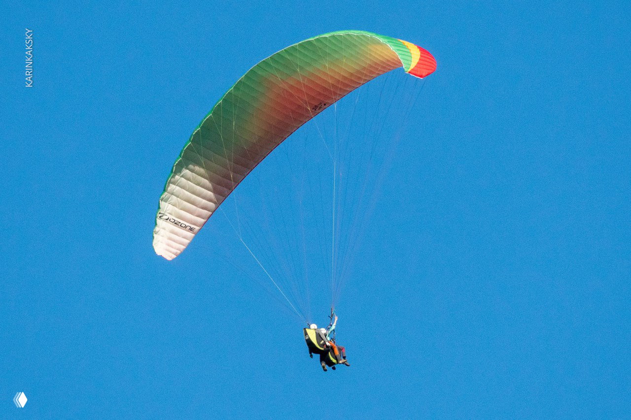 Тандемный полёт крупным планом: купол ярко-зелёный, двое пассажиров в подвеске на фоне чистого голубого неба и лёгкой дымки.