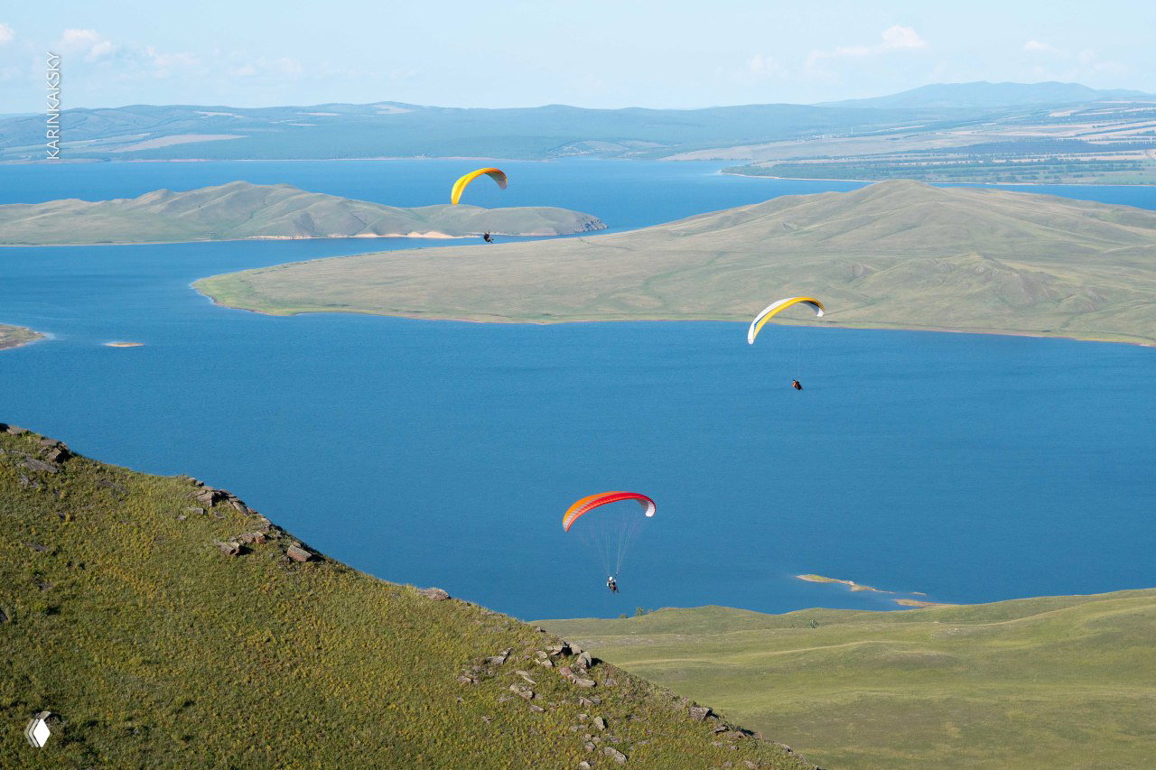 Парапланы в воздухе над большим озером и холмами: несколько экипажей парят над синими водами и зелёными мысами.