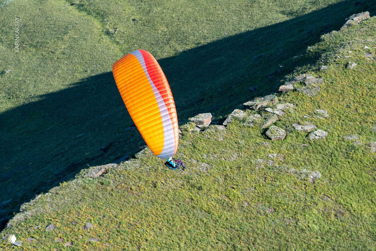 Крупный план оранжевого купола параплана на фоне склона и тени от крылатой формы, пилот в подвесной системе виден частично.