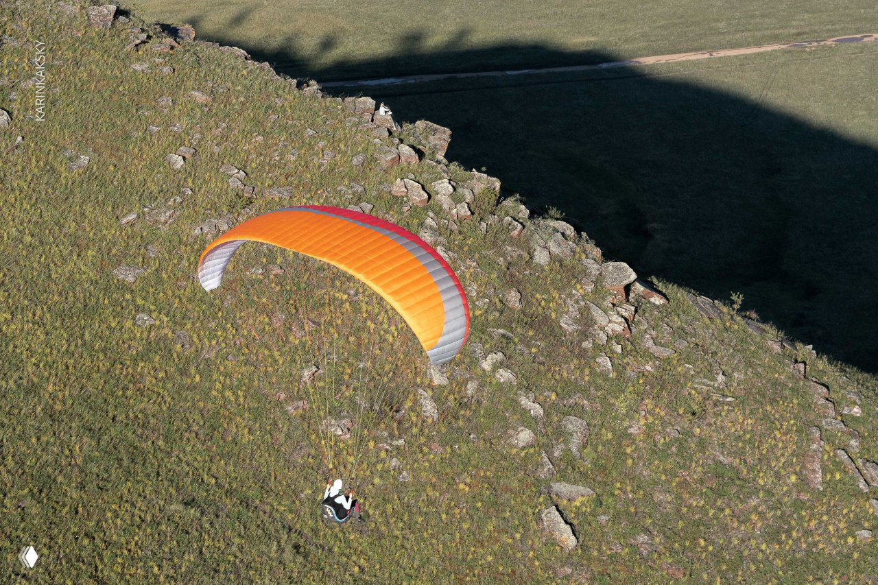 Параплан в полёте по краю скалистого обрыва: вид сверху на купол и травянистый склон, характерная тень от крыла.