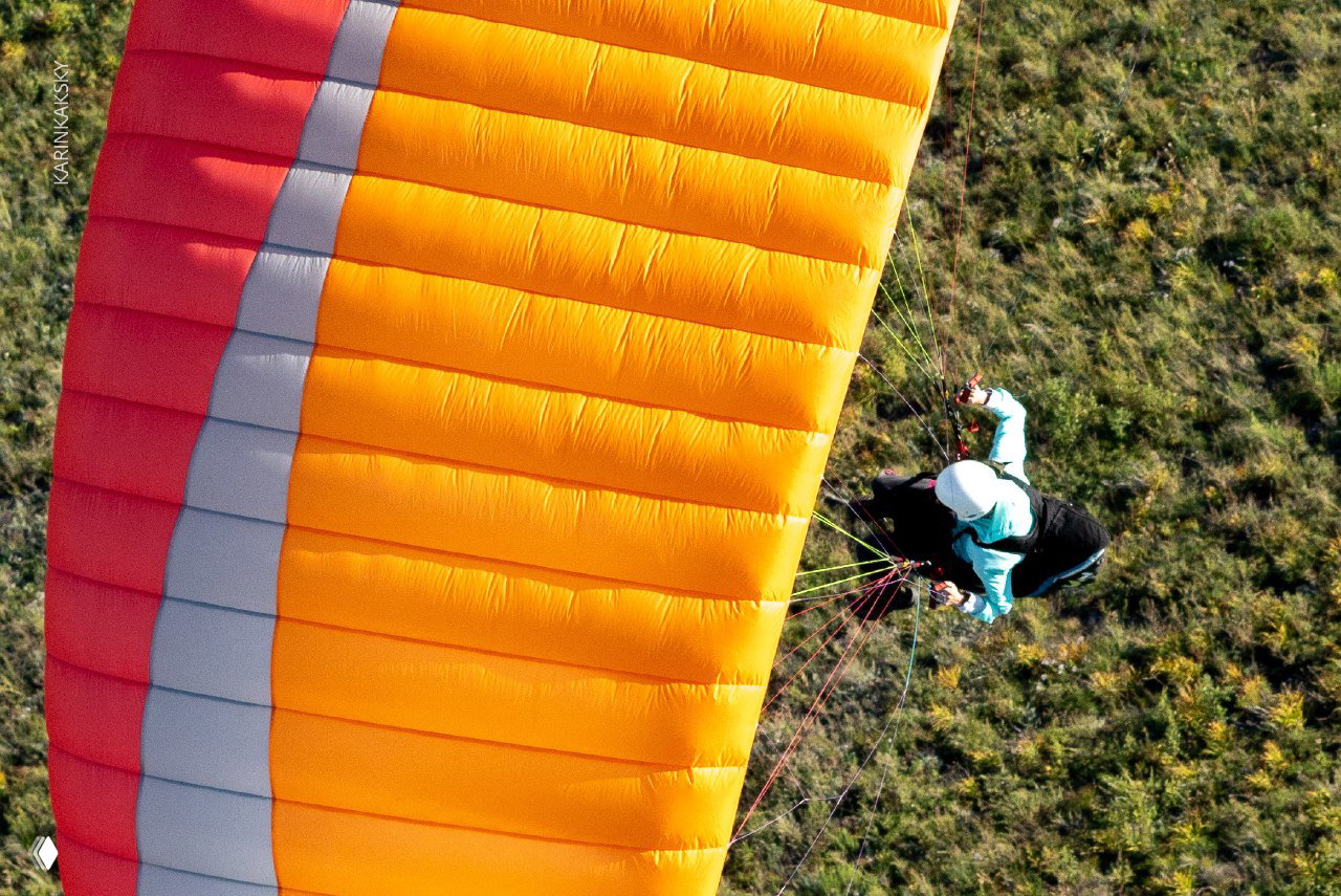 Крупный фрагмент оранжевого параплана в полёте: вид на купол и подвесную систему, пилот манипулирует стропами.