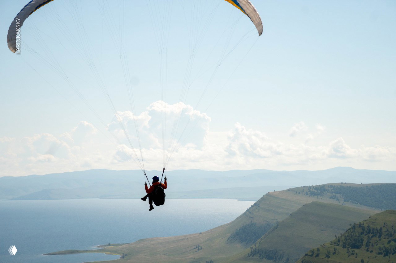 Пилот подпрыгивает на старте: в каске и куртке, на заднем плане зелёные холмы и подготовленные купола на земле.
