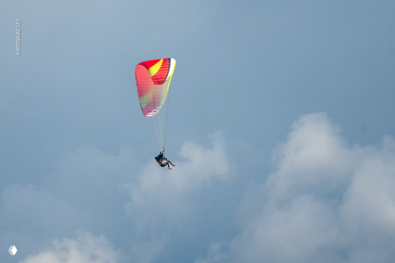 Парапланист в удалённом полёте на фоне облаков: яркий купол и крошечный силуэт пилота в воздушном пространстве.