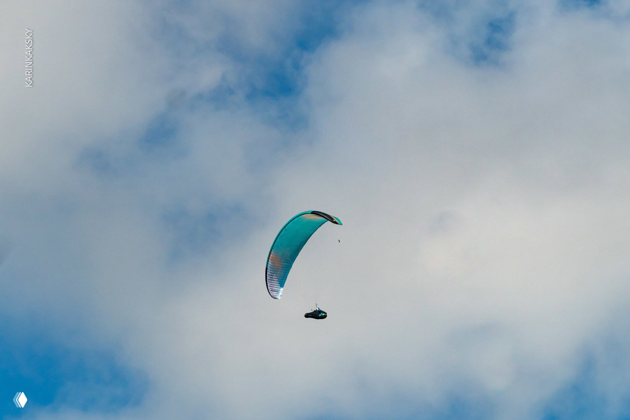 Крупный портрет пилота в синей каске и маске во время подготовки: лицо сосредоточено, на заднем плане полянка и экипировка.