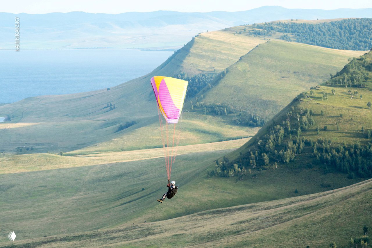 Яркое розово‑жёлтое крыло парящего параплана над холмистой местностью с видом на водоём и удалённые горные гряды.