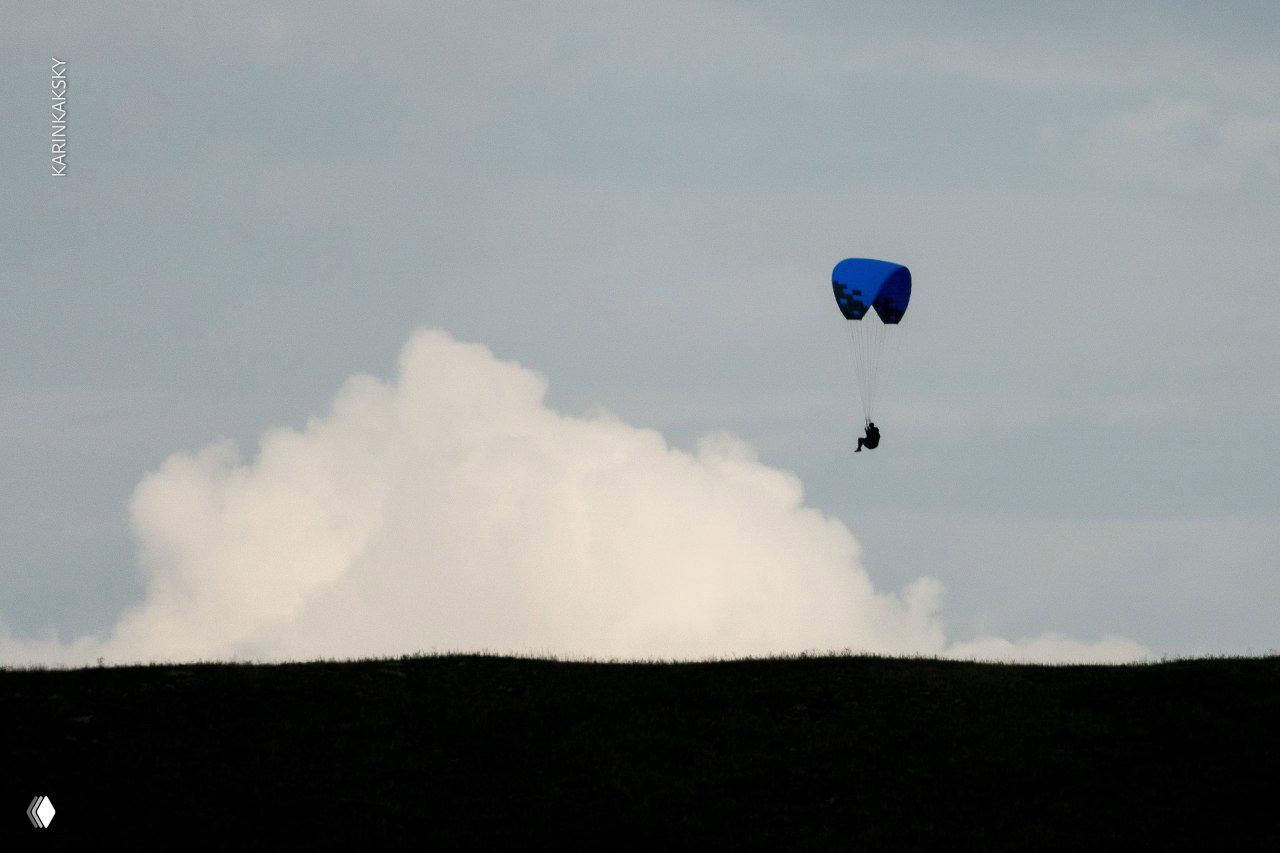 Пилот на синем крыле летит над горизонтом, на заднем плане большое облако и ровная тёмная линия холма.
