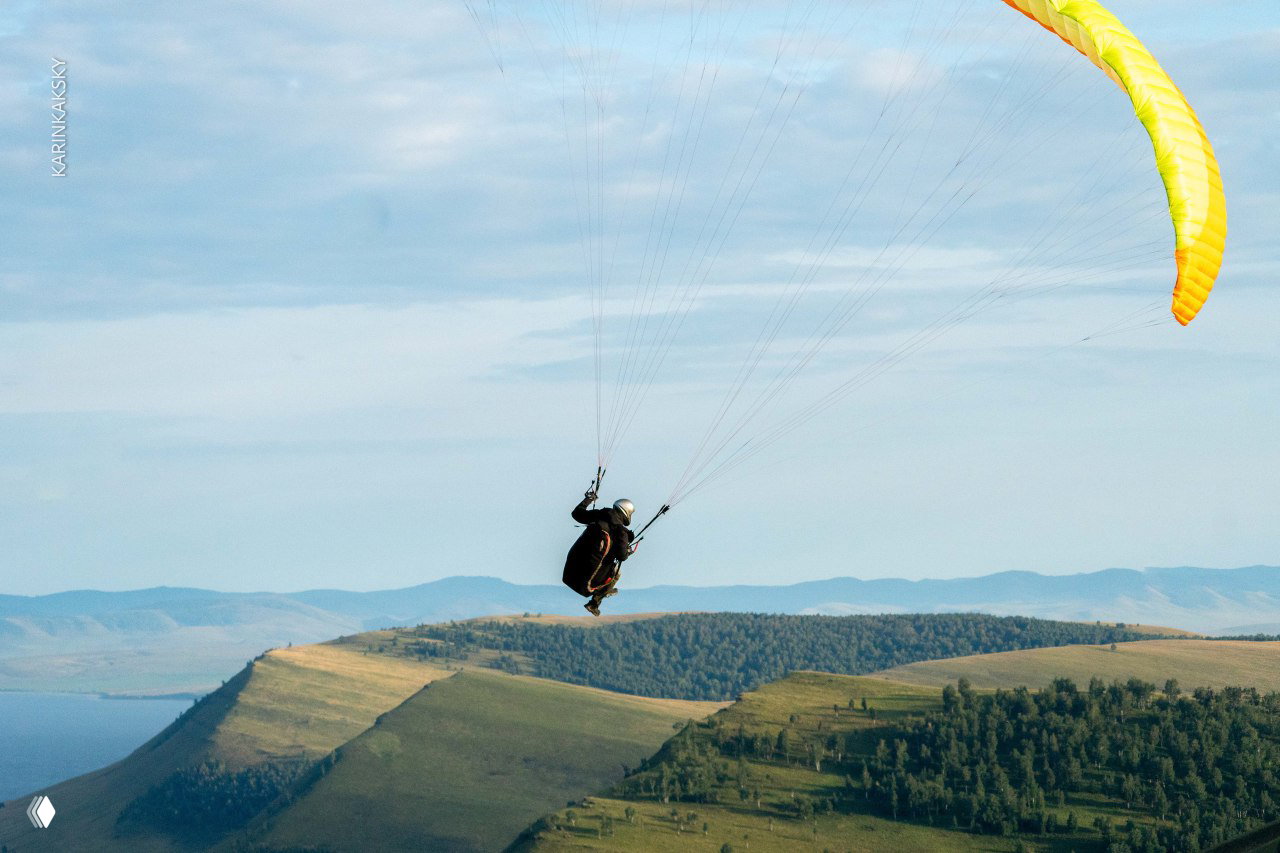 Параплан с жёлтым крылом на фоне холмистой местности и удалённых гор, пилот контролирует крыло в полёте.